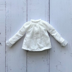 Può includere: Una blusa bianca a maniche lunghe con collo alto e bordi in pizzo sui polsini. La blusa è realizzata in un tessuto leggero e ha un aspetto leggermente drappeggiato.