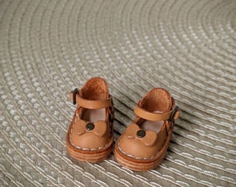 May include: A pair of brown leather doll shoes with a bow and buckle detail. The shoes are on a woven beige surface.