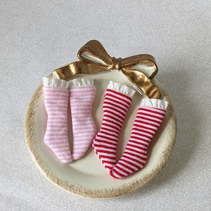 May include: Two pairs of striped doll socks, one pink and white, one red and white, on a white plate with a gold bow.
