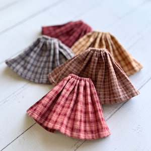 Pu&ograve; includere: Quattro gonne di bambola in tessuto a quadri di vari colori, tra cui rosso, marrone e grigio. Le gonne sono arricciate e hanno un orlo a balze.