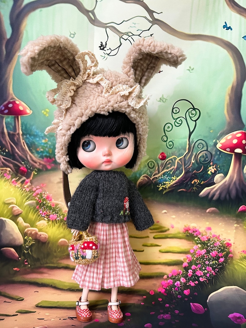May include: A doll wearing a brown bunny hat with white lace trim, a black sweater, and a pink and white gingham skirt. The doll is holding a small basket with mushrooms. The doll is standing on a path in a woodland setting.