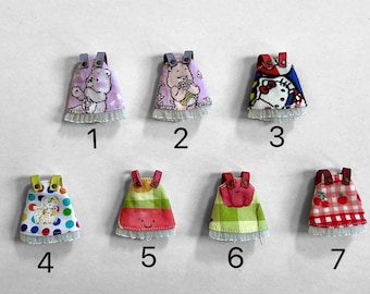 May include: A collection of seven miniature doll dresses. Each dress features a unique design, including Care Bears, Hello Kitty, and fruit patterns. The dresses have a ruffled hem and straps, with the numbers 1 through 7 printed below each dress.