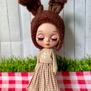 May include: A doll wearing a brown fluffy bunny hat, a tan jumper with a white lamb design, and a brown and white gingham skirt. The doll has pink heart-shaped eye accents and is standing on a red and white checked surface.