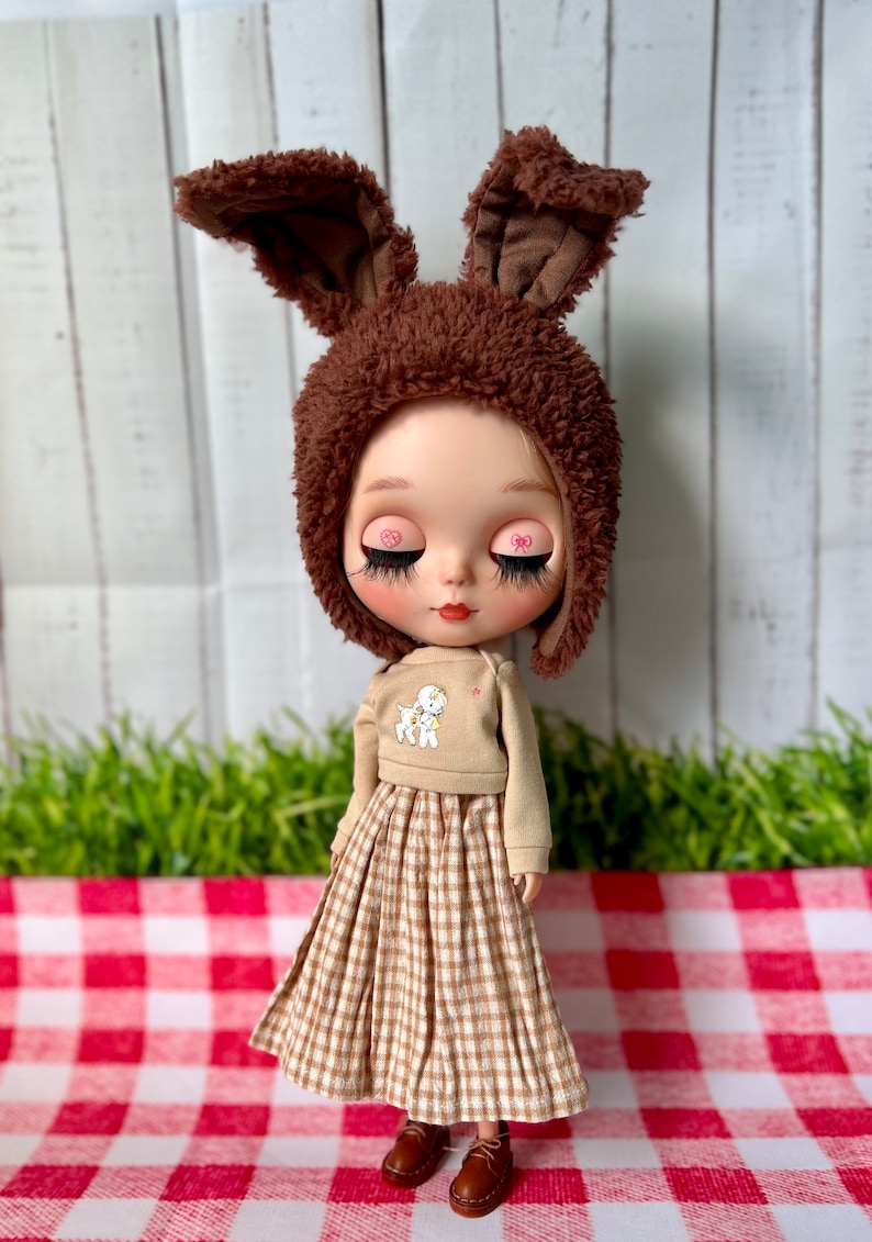 May include: A doll wearing a brown fluffy bunny hat, a tan jumper with a white lamb design, and a brown and white gingham skirt. The doll has pink heart-shaped eye accents and is standing on a red and white checked surface.