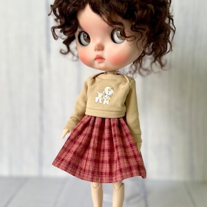 Pu&ograve; includere: Una bambola con capelli castano ricci che indossa un maglione beige con una grafica di un cane bianco e una gonna a quadri rossi e bianchi. La bambola indossa scarpe marroni.