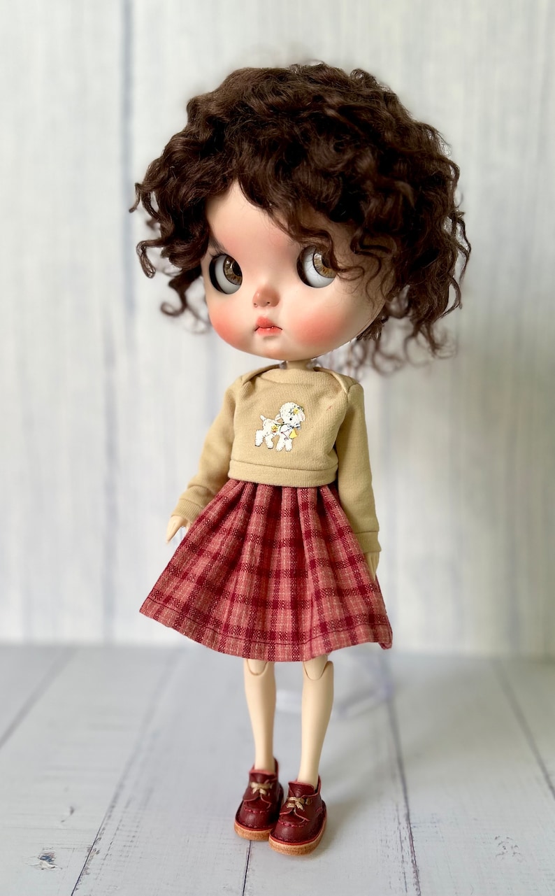 Pu&ograve; includere: Una bambola con capelli castano ricci che indossa un maglione beige con una grafica di un cane bianco e una gonna a quadri rossi e bianchi. La bambola indossa scarpe marroni.