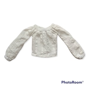 Può includere: Blusa bianca a maniche lunghe con spalle scoperte, scollo arricciato e vestibilità leggermente corta.
