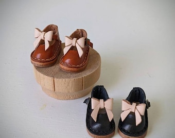 May include: Two pairs of miniature leather shoes with pink bows. The shoes on the left are brown leather and the shoes on the right are black leather.