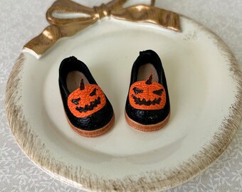 May include: A pair of black doll shoes with orange pumpkin faces. The shoes are on a white plate with a gold bow.