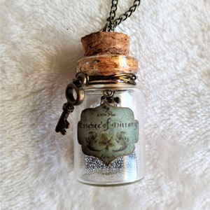 May include: A small glass bottle pendant necklace with a cork stopper and a decorative key charm. The bottle contains small silver beads and a label that reads "Essence of Brittany". The necklace chain is a dark bronze color.