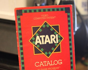 Könnte beinhalten: Ein roter Atari Video Computer System Katalog mit einem grün-schwarzen karierten Rand. Der Katalog zeigt das Atari-Logo und den Text "CATALOG 49 GAME PROGRAM™ CARTRIDGES".