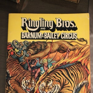May include: A yellow program cover for the Ringling Bros. and Barnum & Bailey Circus. The cover features a lion jumping through a ring of fire, two tigers, and a man in a costume. The text on the cover reads "Ringling Bros. and Barnum & Bailey Circus" and "The Greatest Show on Earth".