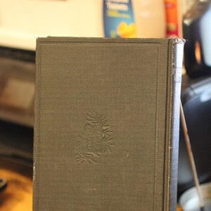May include: A vintage, dark gray book with a textured cover and embossed floral design. The book is displayed on a silver metal stand with small ball feet, set against a blurred background. The book's spine is visible.