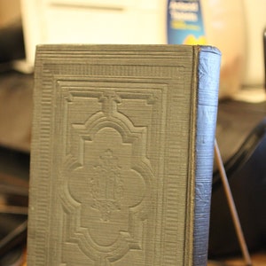 May include: A dark gray hardcover book with an embossed design on the front cover. The design features a central motif with a decorative border.