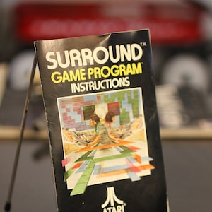 May include: A black and white Atari game program instruction manual for the game Surround. The manual is on a metal stand and features a logo for Atari and the text "SURROUND GAME PROGRAM INSTRUCTIONS TM".