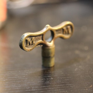 Puede incluir: Una llave de latón vintage con una "M" en cada extremo del mango. La llave tiene una abertura circular en el centro y una base cilíndrica. La llave es probablemente para un mueble o un objeto decorativo.
