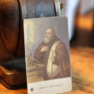 Op de afbeelding: Een vintage religieuze kaart met een illustratie van een heilige met een halo, gekleed in bruine gewaden en een staf vasthoudend. De kaart is geplaatst voor een donkerbruine puntenslijper. De tekst "S. Ignazio da Laconi Cappuccino" is zichtbaar.