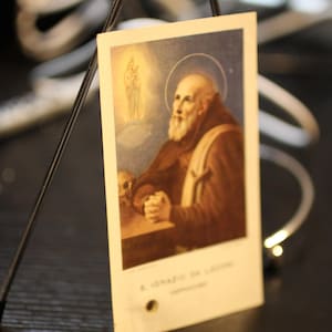 Op de afbeelding: Vintage religieuze kaart met een portret van een heilige met een halo, gekleed in een bruine mantel en een witte baard. De kaart heeft een kleine schedel en tekst aan de onderkant. De achtergrond is zachtblauw en goud.