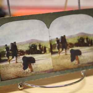 May include: A vintage stereoscopic postcard showing a group of people walking in a line. The postcard is in black and white and has a faded look. The people are wearing clothing from the early 1900s.