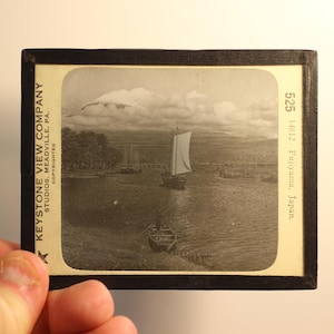 Könnte beinhalten: Ein altes Schwarzweißfoto des Mount Fuji in Japan. Der Berg ist teilweise von Wolken verdeckt. Im Vordergrund ist ein kleines Segelboot zu sehen.