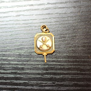 May include: A small, gold-toned pendant with a rectangular frame and a circular center. The center features a design of crossed swords and a figure. The pendant has a loop at the top and a small pin extending from the bottom.