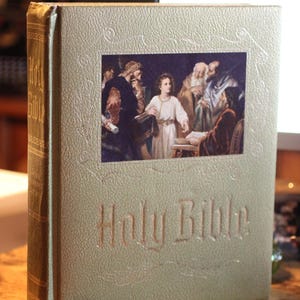 Könnte beinhalten: Eine goldfarbene, gebundene Heilige Bibel mit strukturiertem Einband, geprägtem Rand und dem Schriftzug Holy Bible in Goldbuchstaben. Ein rechteckiges Bild auf der Vorderseite zeigt eine Gruppe von Figuren in historischer Kleidung.
