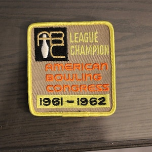 Könnte beinhalten: Ein gestickter Aufnäher in Gelb und Braun mit dem Text "League Champion" und "American Bowling Congress" sowie den Jahren "1961 - 1962". Der Aufnäher zeigt außerdem einen schwarzen und weißen Bowling-Pin.