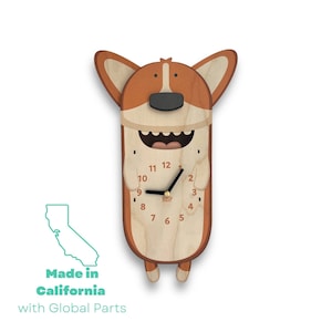 May include: A wooden wall clock shaped like a cartoon corgi dog with a smiling face and a clock face with black hands. The clock is made in California with global parts.