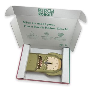 May include: A green wooden clock shaped like a monster with a toothy grin and a clock face with black hands. The clock is in an open box with the words "Birch Robot" printed on the box.