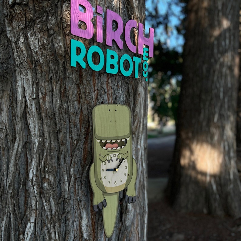 May include: A green wooden dinosaur clock with a white face and black hands hanging on a tree trunk. The clock is labeled "Birch Robot 9".