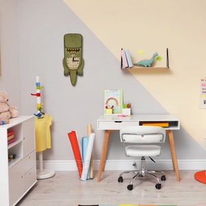 May include: A white desk with a wooden frame and a white chair with wheels. There is a green dinosaur clock on the wall, a shelf with books, and a small dinosaur toy on the shelf.