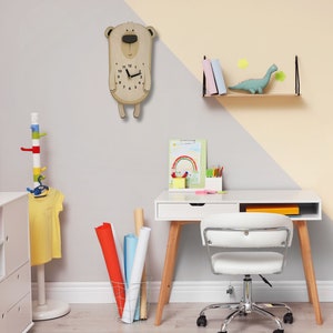 K&ouml;nnte beinhalten: Eine h&ouml;lzerne Uhr in B&auml;renform mit schwarzen Zeigern und Zahlen h&auml;ngt an einer hellgrauen Wand. Im Hintergrund befindet sich ein wei&szlig;er Schreibtisch mit einem wei&szlig;en Stuhl und einem h&ouml;lzernen Dinosaurierspielzeug auf einem Regal.