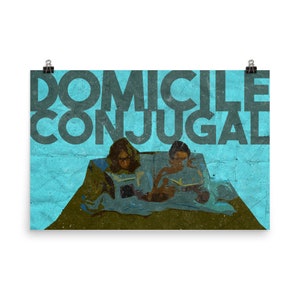 Pode incluir: Pôster com o texto "DOMICILE CONJUGAL" em letras grandes cinza escuro. O pôster apresenta uma ilustração de duas pessoas lendo, com um fundo azul e uma aparência texturizada. A imagem é mantida no lugar por dois clipes pretos.