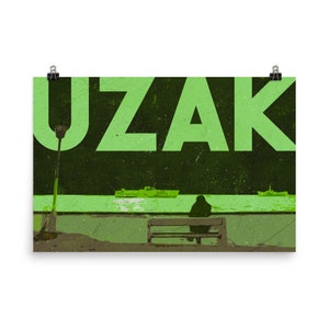 Puede incluir: Un póster verde y negro con la palabra "UZAK" en letras verdes grandes. Una persona se sienta en un banco en primer plano, mirando hacia un cuerpo de agua. El póster tiene un aspecto vintage y desgastado.