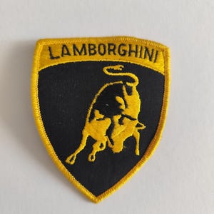 May include: A shield-shaped embroidered patch featuring a black background with a yellow bull silhouette and the text LAMBORGHINI in black letters on a yellow border. The patch has a yellow stitched edge and a pointed bottom design.