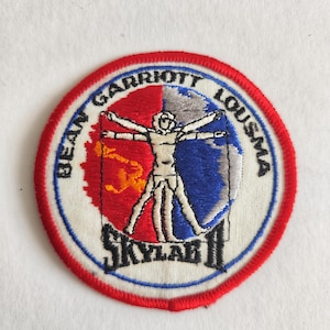May include: A circular embroidered patch featuring a red and blue background with a white Vitruvian Man figure. The text BEAN GARRIOTT LOUSMA is arched across the top, and SKYLAB II is printed in black at the bottom, all bordered by a red edge.