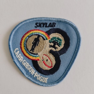 May include: A light blue embroidered patch with a rounded triangular shape. It features the text SKYLAB and CARR-GIBSON-POGUE. The design includes three circular icons: a silhouette with a rainbow, a green tree, and a red-lined oval shape.