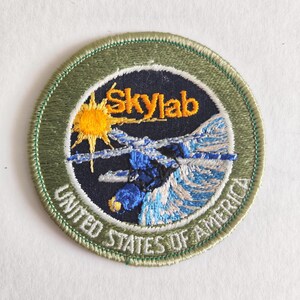 May include: A circular embroidered patch featuring a dark blue background with a yellow sun and the text Skylab in yellow. A blue and white space station illustration is centered, surrounded by a sage green border with United States of America.