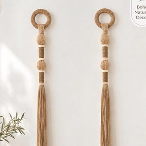 May include: Two identical wall hangings featuring a circular top wrapped in tan jute cord, followed by a series of wrapped spheres and light-colored wooden beads, ending in a long, straight, tan fiber tassel. Text reads Boho Natural Decor.