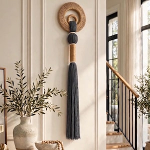 May include: A wall hanging featuring a large tan rope-wrapped ring with a smaller matching ring inside. A dark gray cord hangs from the center, accented by a dark gray sphere, a tan rope-wrapped cylinder, and a long dark gray fringe tassel.