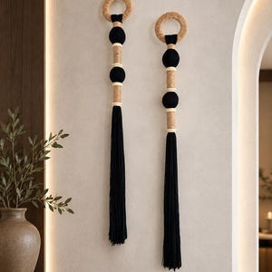 May include: Two vertical wall hangings featuring a top circular ring wrapped in tan jute, followed by alternating black spheres and tan jute-wrapped segments, ending in a long black tassel. The design uses a repeating pattern of natural fiber and black thread.