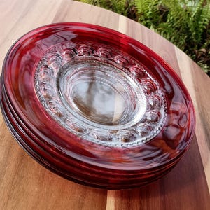 May include: A stack of red glass plates with a clear, textured center. The plates have a decorative, raised pattern around the inner rim. The plates are stacked on a wooden surface.