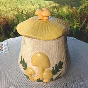 May include: A ceramic mushroom-shaped jar with a lid. The jar is beige with a yellow lid and mushroom accents. The lid has a cluster of small mushrooms as a handle. Green leaves and yellow mushrooms decorate the front of the jar.