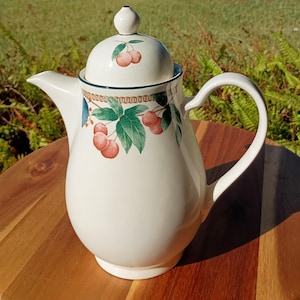 May include: Cream-colored ceramic coffee pot with a lid, handle, and spout. The pot is decorated with a cherry design featuring red cherries and green leaves. The pot sits on a wooden surface with a green background.