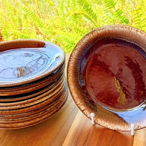 May include: A stack of brown ceramic plates with a glossy finish. The plates have a dark brown rim that fades into a lighter brown. One plate is displayed on a white stand. The plates are on a wooden surface.