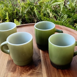 May include: Four vintage green glass mugs with white interiors and rounded handles are arranged on a wooden tray. The mugs have a speckled texture and a gradient color, transitioning from a darker green at the base to a lighter green.
