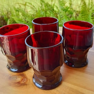 May include: Four ruby red drinking glasses with a faceted design. The glasses are made of a transparent material and have a cylindrical shape with a wider base. The glasses are arranged on a wooden surface.