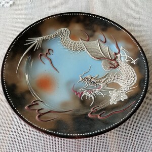 May include: A round decorative plate featuring a central light blue background with a gradient transition to a dark brown border. A raised, textured silver dragon motif curves across the surface, accented with small red and white painted details.