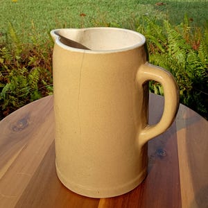 May include: A tan ceramic pitcher with a large handle and a spout. The pitcher has a cylindrical body and a light-colored interior. It is sitting on a wooden surface.