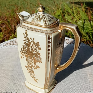 May include: Cream-colored porcelain teapot with gold floral designs and accents. The teapot has a square shape with a curved handle and a lid with a gold finial. The floral design is repeated on the lid and body.